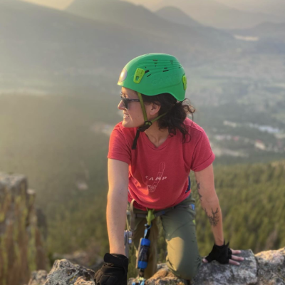 Woman rock climbing in Estes Park with red shirt and green helmet looking off into the distance with valley view behind her
